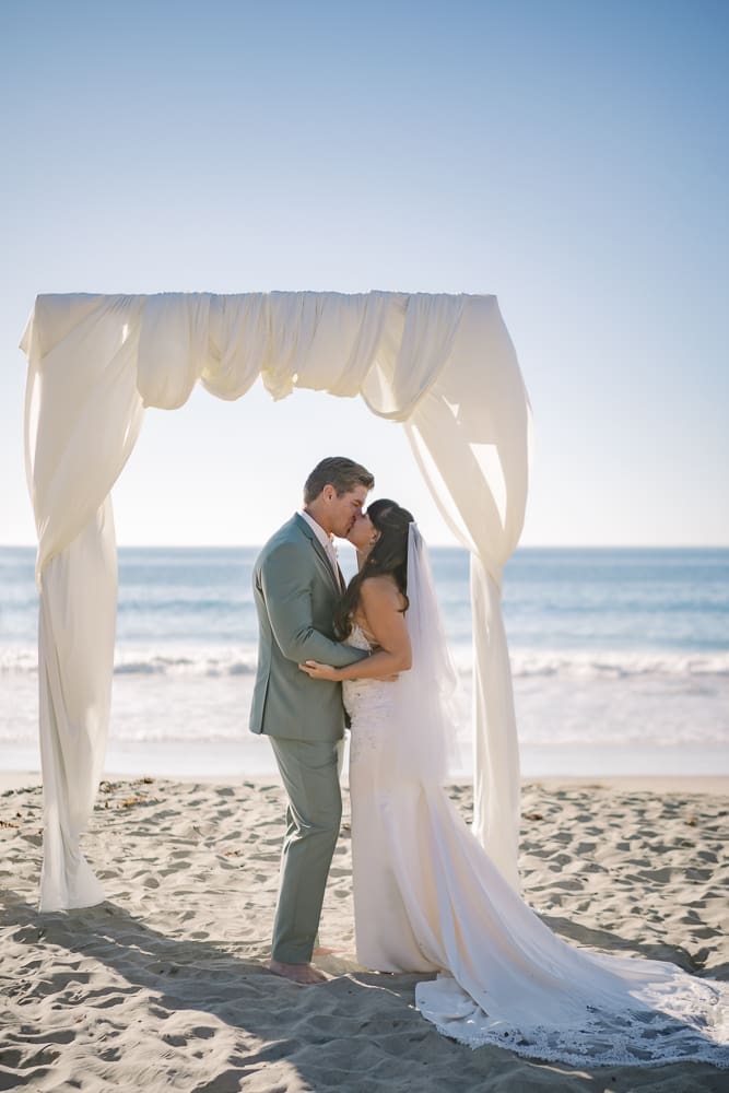 Bride and groom kissing under white drapery arch on Torrance Beach